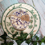 Medium Wild Flowers soy candle 500g with blue cornflowers decoration on white wax, styled with fresh ivy vines on vintage leaf-patterned ceramic plate, rustic whitewashed wood background