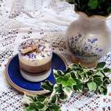 Candle with decorative elements on a lace tablecloth with a vase and plants.