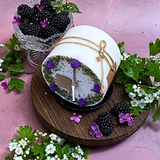 Decorative candle on a wooden board with blackberries and flowers.