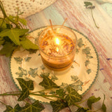 Small Glass Wild Flowers soy candle 400g burning with golden flame glow in clear glass container, blue cornflowers visible in melted wax pool, styled on ivy-patterned plate with fresh ivy - glass container candle burning demonstration photography