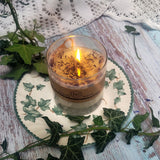 Small Glass Wild Flowers soy candle 400g burning with lit flame and Beauty Scents label in clear glass container, blue cornflowers floating in melted wax pool, styled on ivy-patterned plate with fresh ivy and white lace - glass container burning ambiance photography