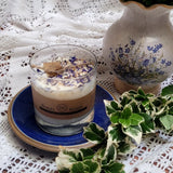 Small Glass Wild Flowers soy candle 400g in clear glass container with blue cornflowers decoration on blue ceramic plate, styled with white lace doilies, floral vase and fresh variegated foliage - glass container lifestyle photography