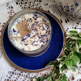 Small Glass Wild Flowers soy candle 400g in clear glass container with blue cornflowers decoration on blue ceramic plate, styled with white lace doilies, floral vase and fresh variegated foliage - glass container lifestyle photography