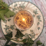 Small Glass Wild Flowers soy candle 400g burning with golden flame glow in clear glass container, blue cornflowers visible in melted wax pool, styled on ivy-patterned plate with fresh ivy - glass container candle burning demonstration photography