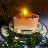 Small Heart Wild Flowers soy candle 300g burning with lit flame and melted wax pool, blue cornflowers decoration visible in heart shape, styled on ivy-patterned plate with fresh ivy - heart-shaped candle burning demonstration photography