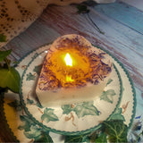 Small Heart Wild Flowers soy candle 300g burning with golden flame glow in heart-shaped wax pool, blue cornflowers visible around edges, styled on ivy-patterned plate with fresh ivy - romantic heart candle burning ambiance photography