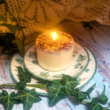 Small wild flowers candle with blue cornflowers burning with warm golden flame on vintage ivy patterned china plate with fresh greenery