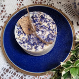 Small handmade wild flowers soy candle with blue cornflower petals styled on ceramic plate in rustic home decor setting