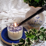 Small wild flowers candle with blue cornflowers being lit with long lighter on blue ceramic plate with vintage floral pot and lace