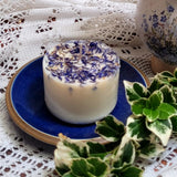 Small wild flowers scented candle with blue cornflowers on elegant blue ceramic plate styled with white lace and fresh green foliage